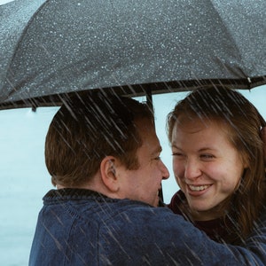 May include: A couple standing under a black umbrella in the rain, smiling and looking at each other.