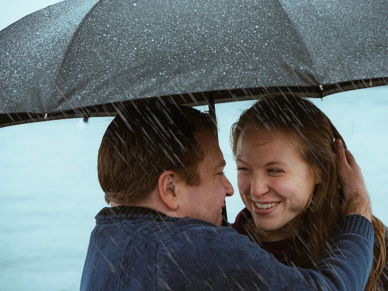 May include: A couple standing under a black umbrella in the rain, smiling and looking at each other.