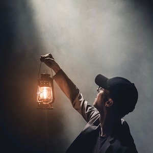 May include: A person wearing a black cap and glasses holds a lit kerosene lantern in a smoky room.