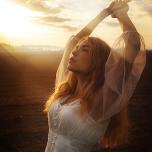 May include: A woman with long, blonde hair wearing a white, sheer dress with lace detailing. She is standing in a field with her arms raised above her head, looking up at the sunset.