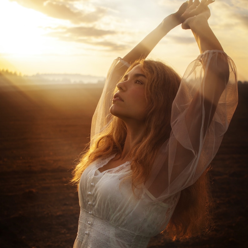 May include: A woman with long, blonde hair wearing a white, sheer dress with lace detailing. She is standing in a field with her arms raised above her head, looking up at the sunset.