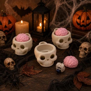 May include: Three off-white skull-shaped keycap holders, two containing pink brain-shaped keycaps, with one additional pink brain keycap and a mechanical keyboard switch placed on a dark surface among decorative Halloween items.
