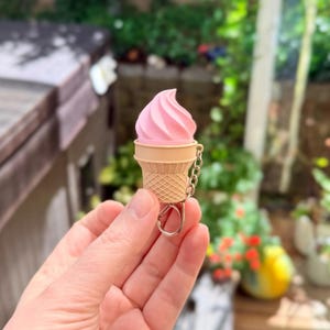 May include: A keychain featuring a miniature soft-serve ice cream cone with a light pink swirled top and a textured tan waffle cone base. The accessory includes a silver-toned metal chain and a circular ring for attachment to keys or bags.