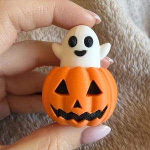 May include: A small, three-dimensional object featuring a white ghost with black eyes and a mouth, emerging from the top of a bright orange jack-o'-lantern pumpkin. The pumpkin has carved black triangular eyes and a jagged black mouth.