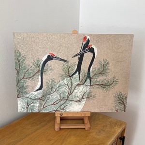Op de afbeelding: Een vintage-stijl illustratie van drie roodgekroonde kraanvogels met zwart-wit verenkleed, die op een tak van een dennenboom zitten. De achtergrond is een beige canvas met een delicaat bloemenpatroon.