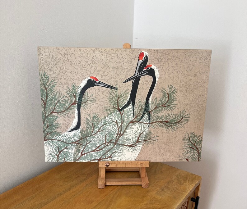 Op de afbeelding: Een vintage-stijl illustratie van drie roodgekroonde kraanvogels met zwart-wit verenkleed, die op een tak van een dennenboom zitten. De achtergrond is een beige canvas met een delicaat bloemenpatroon.