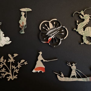 Op de afbeelding: Een verzameling van acht papieren uitsnijdingen met verschillende ontwerpen, waaronder een draak, een bloem, een persoon met een paraplu, een persoon met een bloem, een persoon met een zwaard, een persoon in een boot en een plant. De uitsnijdingen zijn gemaakt van gepatroneerd papier in de kleuren groen, wit, zwart en rood.