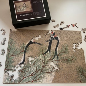 Op de afbeelding: Een 1000-delige puzzel met een gedetailleerde illustratie van twee kraanvogels in een dennenboom. De puzzel is gemaakt door Momoyogusa en heet "Kamikura Sanko".