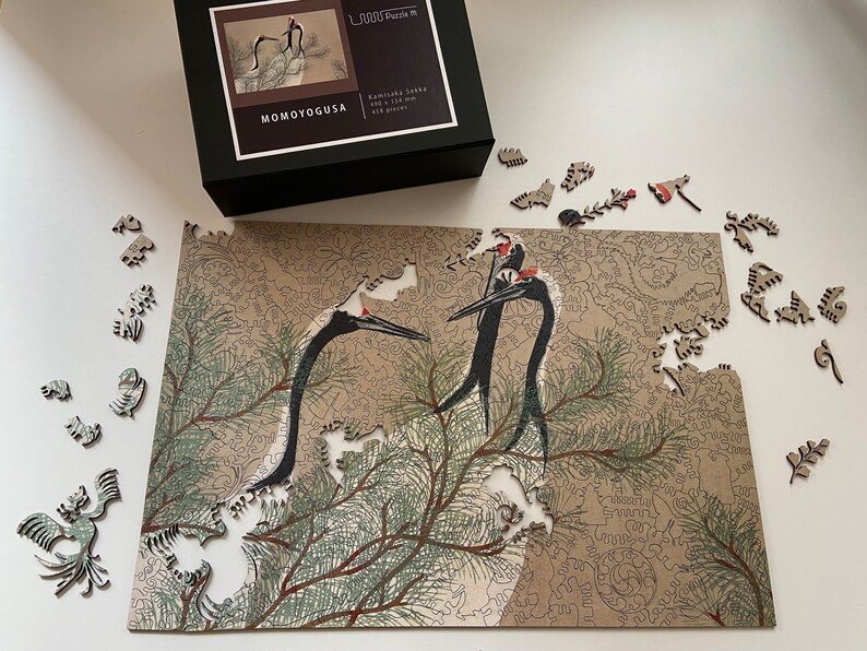 Op de afbeelding: Een 1000-delige puzzel met een gedetailleerde illustratie van twee kraanvogels in een dennenboom. De puzzel is gemaakt door Momoyogusa en heet "Kamikura Sanko".