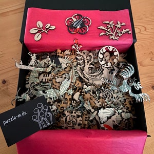 Op de afbeelding: Een zwarte doos met een rode voering en een verscheidenheid aan houten puzzelstukjes erin. De stukjes zijn in de vorm van bloemen, bladeren en andere ontwerpen. De doos bevat ook een klein zwart kaartje met de tekst "puzzle-m.de".