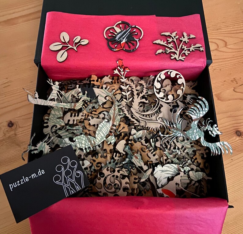 Op de afbeelding: Een zwarte doos met een rode voering en een verscheidenheid aan houten puzzelstukjes erin. De stukjes zijn in de vorm van bloemen, bladeren en andere ontwerpen. De doos bevat ook een klein zwart kaartje met de tekst "puzzle-m.de".