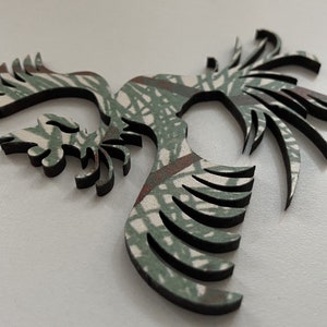 Op de afbeelding: Een houten uitsnijding van een gestileerde vogel met uitgespreide vleugels, in een camouflagepatroon van groen en bruin.