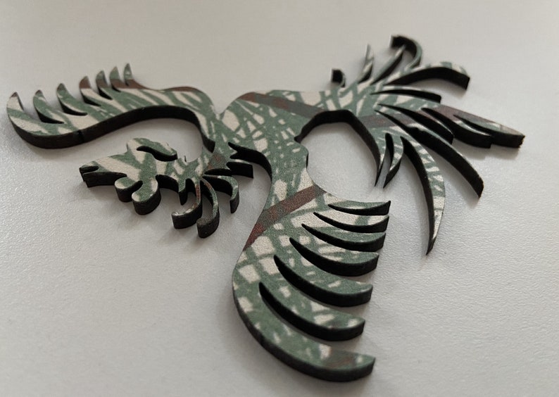 Op de afbeelding: Een houten uitsnijding van een gestileerde vogel met uitgespreide vleugels, in een camouflagepatroon van groen en bruin.