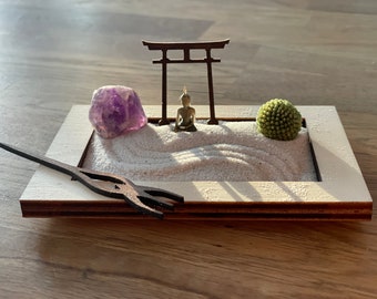 May include: A small wooden zen garden with a white sand base, a miniature Buddha statue, a purple amethyst crystal, a small green bush, and a wooden rake. The garden is framed in wood and has a small wooden torii gate.