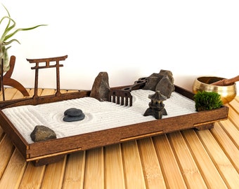 May include: A wooden zen garden tray with white sand, grey stones, a small wooden bridge, a miniature pagoda, and a rake. The tray is on a bamboo mat.