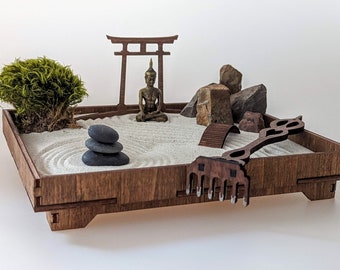 May include: A wooden zen garden with a miniature Buddha statue, a small green bush, a wooden archway, and a rake. The garden is filled with white sand and has a small bridge and a pile of rocks.