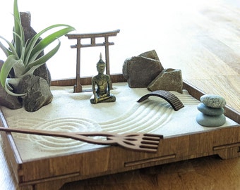 Op de afbeelding: Een houten zen-tuin met een miniatuur Boeddhabeeld, een kleine brug en een hark. De tuin is gevuld met wit zand en heeft een kleine groene plant in de hoek.