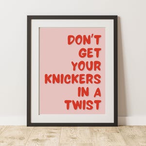 May include: A framed print with a black frame and white matting. The print features the phrase "DON'T GET YOUR KNICKERS IN A TWIST" in red letters on a pink background. The print is displayed on a wooden floor.
