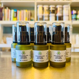 May include: A collection of green glass bottles with black pumps and clear caps. Each bottle has a white label with the text "HYALURONIC ACID ALOE VERA FACE SERUM" and "Wt. 1 oz (28 gms)". The bottles are arranged on a reflective surface.