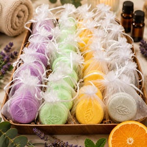 May include: A collection of round, disc-shaped bath bombs in purple, green, yellow, and white, each individually packaged in a sheer white organza drawstring bag. Each bath bomb features a stamped leaf design on the front surface.