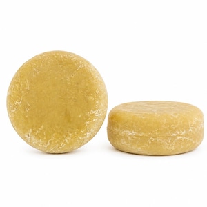May include: Two round, solid shampoo bars with a textured, golden-yellow surface. One bar is positioned upright to show its circular face, while the other rests on its side, displaying its cylindrical thickness and consistent, matte finish.
