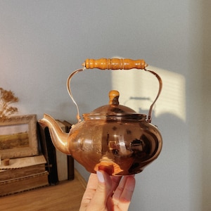May include: A copper teapot with a wooden handle. The teapot is shiny and has a rounded body.