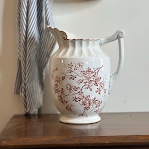 May include: A white ceramic pitcher with a brown floral pattern. The pitcher has a fluted body and a handle with a decorative scroll design.