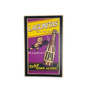 May include: A vintage-style wooden sign advertising Lagunitas India Pale Ale. The sign features a cartoon character boxing a bottle of beer with the text "It's a knock out!" and "Take some along!"