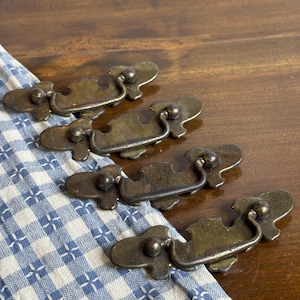 Könnte beinhalten: Vier antike Messing-Schubladengriffe mit dekorativem Design. Die Griffe haben einen gebogenen Griff und einen abgerundeten Knopf an jedem Ende.