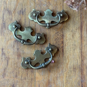 May include: Three antique brass drawer pulls with a decorative, ornate design. Each pull features a curved handle and a detailed backplate with floral-like accents. The pulls are set against a warm, wooden background.