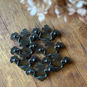 May include: A collection of antique bronze drawer pulls arranged on a wooden surface. Each pull features a decorative design with a curved handle and ornate detailing. The pulls are likely for furniture restoration or vintage decor.