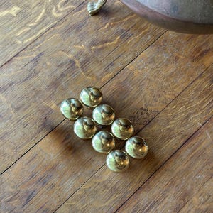 May include: A collection of nine shiny, gold-colored cabinet knobs arranged on a wooden surface. The knobs are spherical with a reflective surface. A portion of a copper-colored pot with decorative feet is visible in the background.