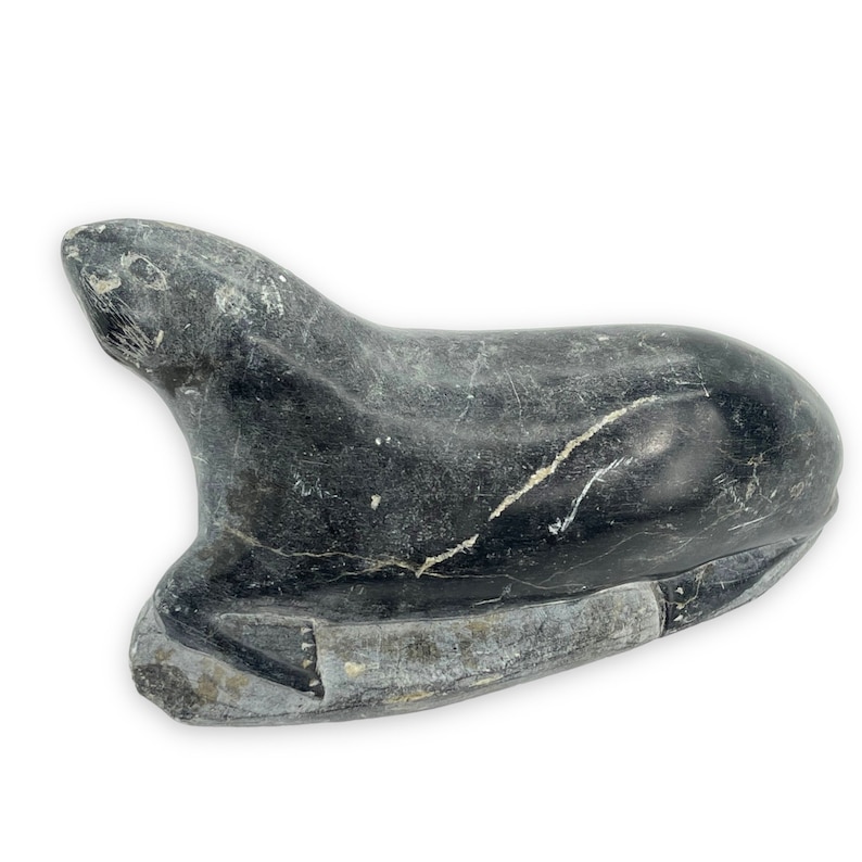 May include: A black and grey carved stone sculpture of a seal lying on its belly. The sculpture is made of a smooth, polished stone with visible veins.