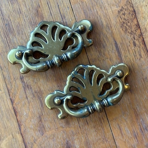 以下が含まれることがあります： 装飾的な透かし彫りデザインの、真鍮製のアンティーク引き出し取っ手2つ。各取っ手は、曲がったハンドルと装飾的なディテールが特徴で、家具にヴィンテージ感を加えるのに最適です。取っ手は木製の表面に置かれています。