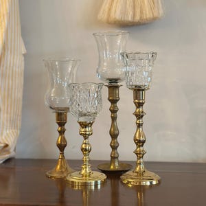 May include: Four vintage brass candle holders with clear glass cups. The candle holders have ornate bases and stems, with varying designs. The glass cups are different shapes and textures, adding visual interest to the arrangement.