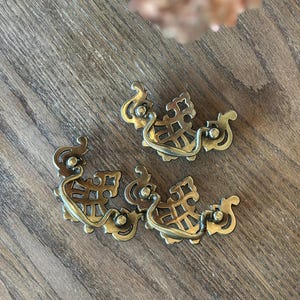 Könnte beinhalten: Vier antike Schubladengriffe aus Messing mit kunstvollen Schnörkeln und einem dekorativen Gittermuster. Die Griffe sind auf einer Holzoberfläche angeordnet und zeigen ihre Vintage-Ästhetik. Jeder Griff hat einen gebogenen Griff und aufwendige Details.