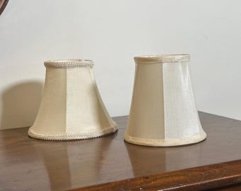 May include: Two off-white lampshades with a pleated design. The lampshades are sitting on a wooden surface.