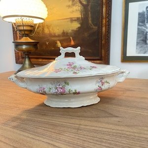 May include: A white porcelain serving dish with a floral pattern in shades of pink and green. The dish has a lid with a handle and a scalloped edge.