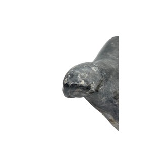 May include: A carved stone sculpture of a seal, with a gray and white color scheme. The seal is lying on its side, with its head turned to the side. The sculpture is made of a smooth, polished stone.