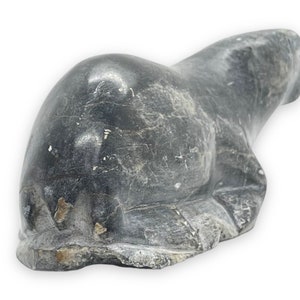 May include: A hand-carved stone sculpture of a seal, lying on its belly with its flippers tucked in. The sculpture is made of dark grey stone with a smooth, polished finish.