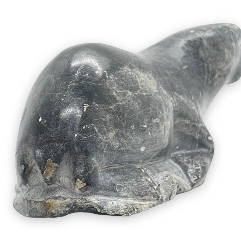 May include: A hand-carved stone sculpture of a seal, lying on its belly with its flippers tucked in. The sculpture is made of dark grey stone with a smooth, polished finish.