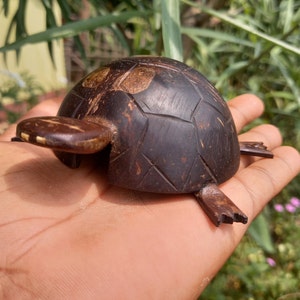 Pode incluir: Uma pequena figura de tartaruga de madeira esculpida à mão com um acabamento marrom escuro. A tartaruga é feita de uma casca de coco e tem um padrão de casca detalhado.