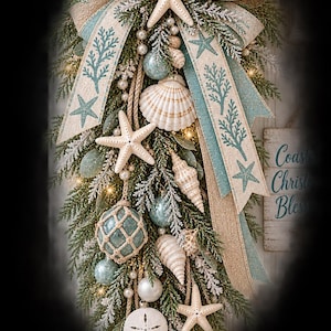 Puede incluir: Un adorno en forma de l&aacute;grima con tem&aacute;tica costera que incluye ramas de pino nevadas, cinta azul claro y arpillera con estampados de coral y estrellas de mar, estrellas de mar blancas, conchas, perlas y adornos de cristal azul claro.