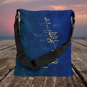 May include: A blue canvas tote bag with a map of the Outer Banks, North Carolina. The map features the names of towns and landmarks, including Corolla, Kitty Hawk, Kill Devil Hills, and Nags Head. The bag has a black adjustable strap.