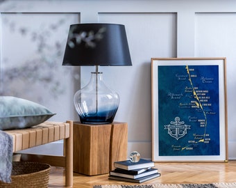 May include: A framed map of the Outer Banks, North Carolina, with a gold anchor and the text "Outer Banks NC" in a gold circle. The map is on a blue background with gold lettering and lines. The map is hanging on a white wall above a wooden table with a lamp and a stack of books.