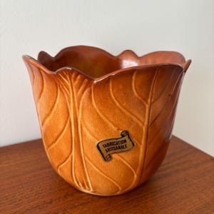 Peut inclure: Un pot de fleurs en céramique avec un motif de feuille dans les tons marron et orange. Le pot a un bord festonné et une étiquette qui dit "FABRICATION ARTISANALE". Cette pièce décorative convient à l'intérieur ou à l'extérieur.