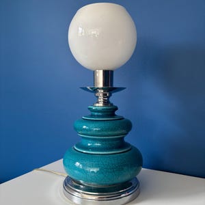Peut inclure: Une lampe de table en céramique turquoise avec un abat-jour globe blanc. La lampe a une finition craquelée et une base et des accents en métal argenté. La lampe est sur une surface blanche contre un mur bleu.