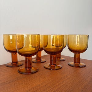 Peut inclure: Ensemble de six verres à pied ambrés. Chaque verre présente une coupe arrondie et un pied court. Ils sont disposés sur une surface en bois. Les verres ont une couleur chaude et un design vintage.