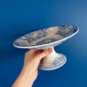 Puede incluir: Un soporte para pasteles de cerámica blanca y azul con un diseño floral. El soporte tiene un plato ancho y poco profundo sostenido por una base de pedestal. El patrón floral azul se repite en la base y el plato. El fondo es azul liso.