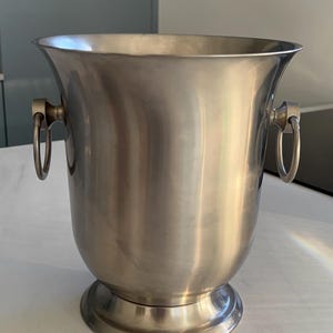 Peut inclure: Seau à vin en acier inoxydable avec un dessus évasé et une base circulaire. Le seau a deux poignées en anneau de chaque côté. Le métal poli reflète la lumière, créant des reflets et des ombres.